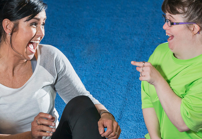 Two women, one with Down’s syndrome, sit on a blue floor, laughing and smiling at each other. One wears a grey shirt, the other a bright green shirt, and they appear to be sharing a joyful moment.