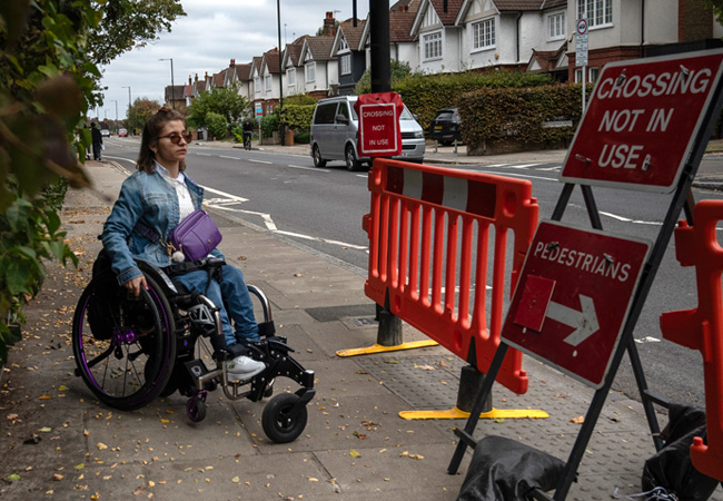 A woman in a wheelchair faces a blocked pavement with barriers and a sign that reads Crossing Not In Use, directing pedestrians to cross elsewhere along a residential street.