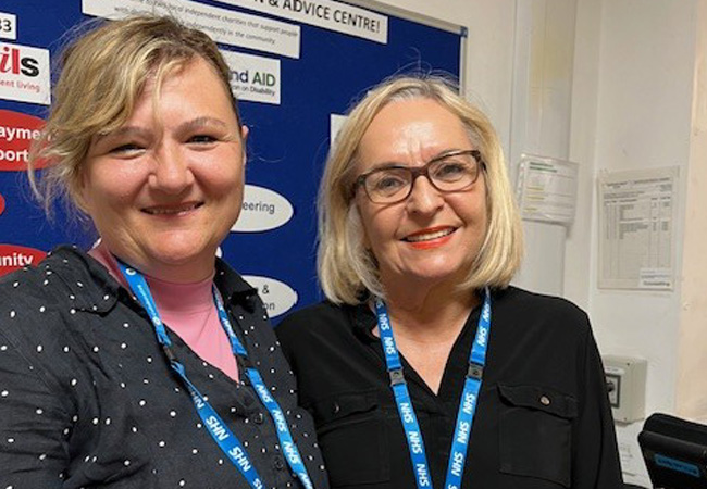 Two women wearing NHS lanyards stand close together and smile at the camera. They are indoors, with a blue information board and documents posted on the wall behind them.