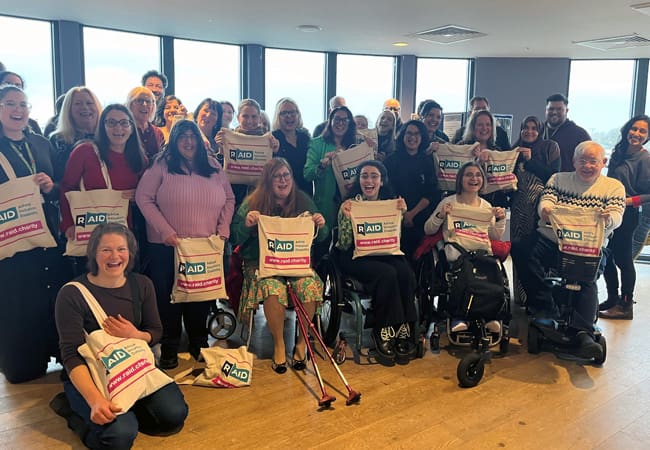 A diverse group of people, including wheelchair users, smile and hold tote bags with “Raid” and “Remove All Barriers” printed on them, gathered in a bright room with large windows.