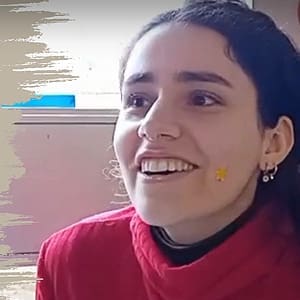 A young woman with dark hair, wearing a red polo-neck jumper, smiles brightly. She has a small yellow flower sticker on her cheek and is indoors with a blurred background.