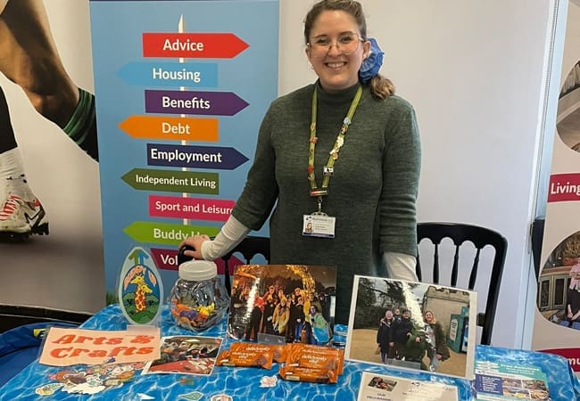 A woman stands smiling behind a table with leaflets, photos, and snacks, at a community event. Behind her is a colourful sign listing services such as advice, housing, benefits, debt, and employment.