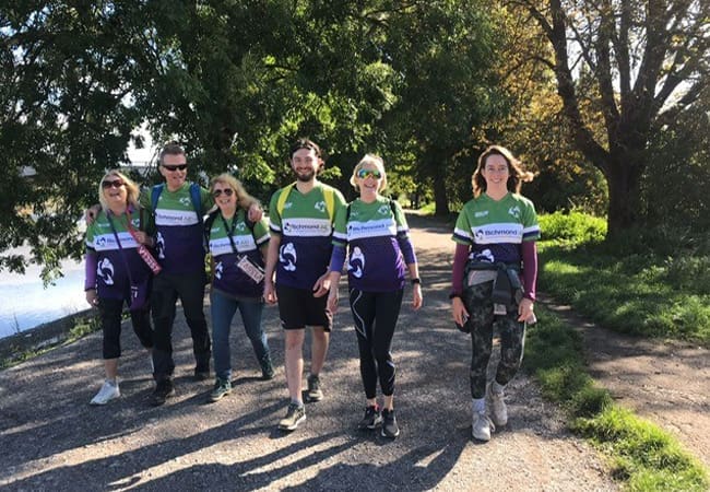 Six people walk together on a sunny path by trees and water, all wearing matching green and purple shirts with white stripes and logos. They appear cheerful and ready for an outdoor event.