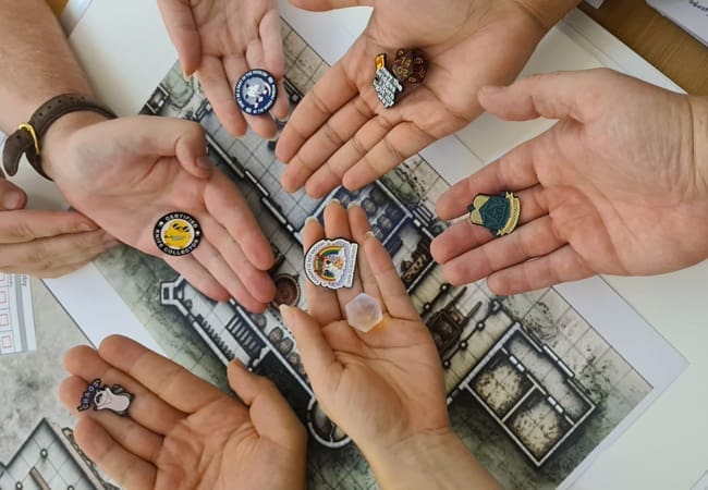 Six hands hold colourful game tokens over a tabletop map, ready for a thrilling dungeon RAID. The map’s rooms and corridors set the stage for an adventure—perfect inspiration for our September Newsletter!.