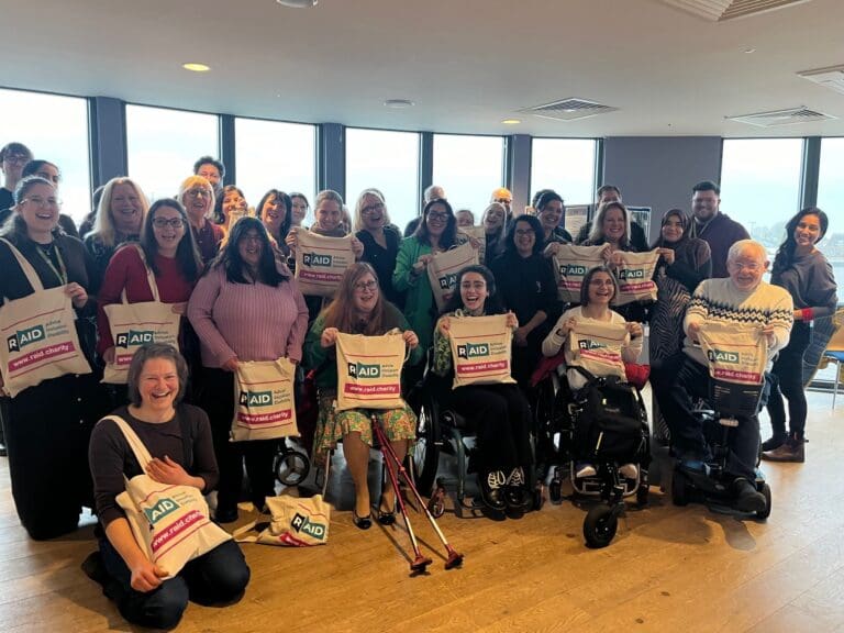 Photo of a group of people, some sitting and some standing, holding white tote bags with the new RAID ( Richmond AID ) logo. Announcing and celebrating the rebranding of Richmond AID to RAID.