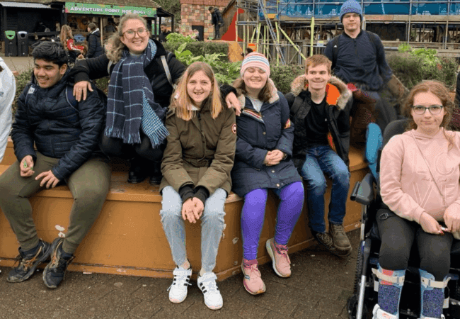 A group of seven young people, some smiling, sit together outdoors on a low wall in Kingston. One person uses a wheelchair. They wear winter clothes and appear to be at a park with scaffolding and plants in the background.