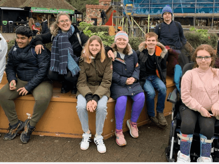 A group of seven young people, some smiling, sit together outdoors on a low wall in Kingston. One person uses a wheelchair. They wear winter clothes and appear to be at a park with scaffolding and plants in the background.