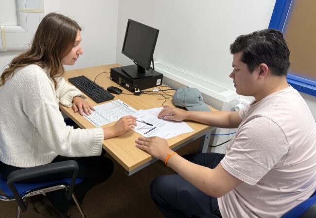 Picture of two people seated at a desk containing a computer. There are documents on the desk which the people are discussing.