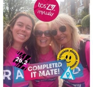 Three smiling women in pink event shirts stand together outdoors, proudly representing the RAID Walkers. Overlaid stickers read “26.2,” “COMPLETED IT MATE!,” “FINISHER,” and “tcs myway 2026.” Houses and sunny sky fill the background.