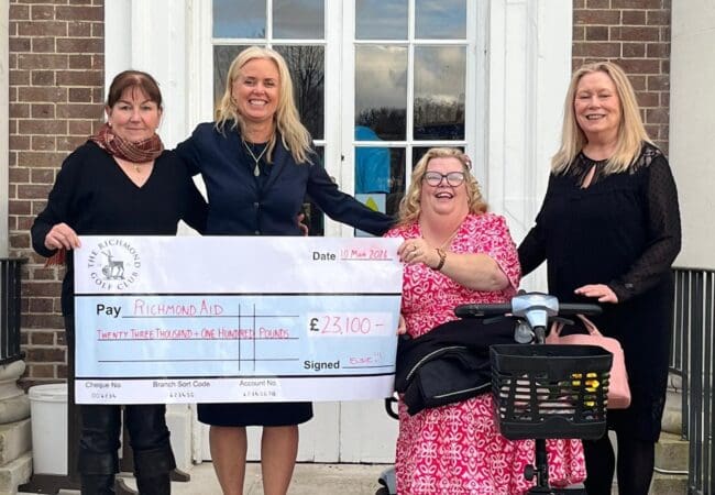 Four women smiling and posing outside a brick building, holding a large cheque for £23,100 made out to Richmond AID. One woman is seated in a wheelchair