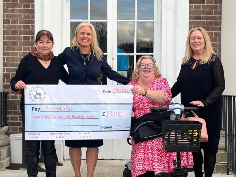 Four women smiling and posing outside a brick building, holding a large cheque for £23,100 made out to Richmond AID. One woman is seated in a wheelchair