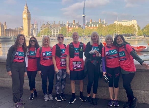 A group of eight RAID Walkers wearing matching shirts stand smiling by the River Thames, with Big Ben and the Houses of Parliament visible in the background on a sunny day in London.