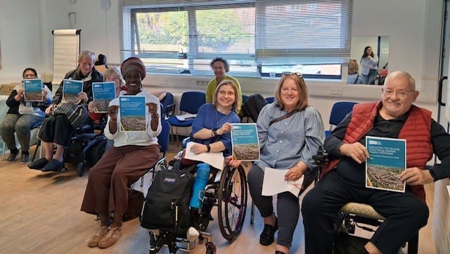 A group of people, some in wheelchairs, sit in a bright room holding up the RAID Housing Report and smiling at the camera.