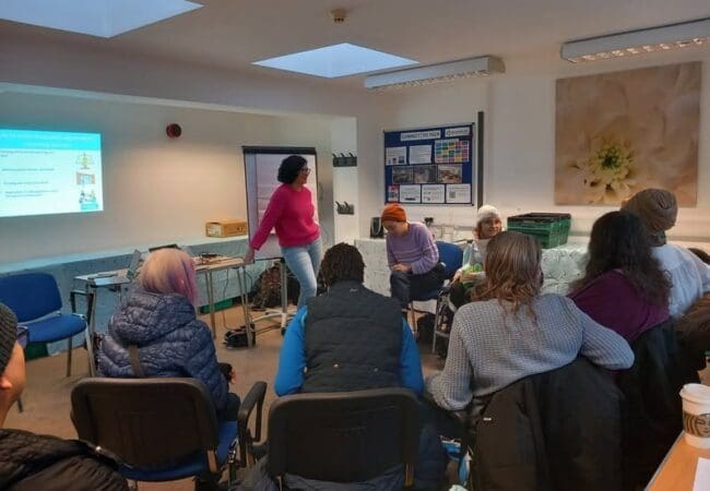 RAID Housing Workshop A group of people sit in a semi-circle in a brightly lit room, listening as a woman presents slides from the RAID Housing Report: Crisis Facing Disabled People in Richmond. The atmosphere appears informal and engaging.