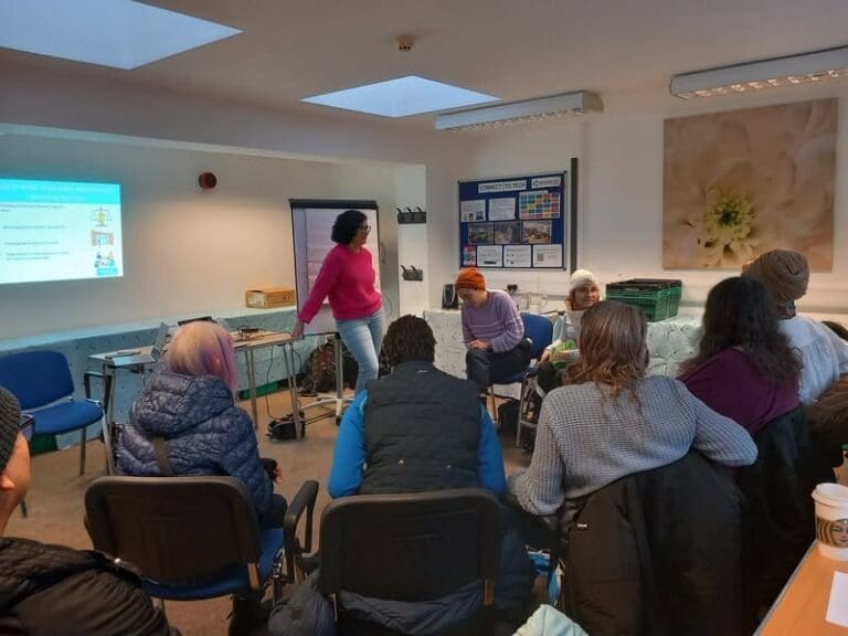 A group of people sit in a semi-circle in a brightly lit room, listening as a woman presents slides from the RAID Housing Report: Crisis Facing Disabled People in Richmond. The atmosphere appears informal and engaging.