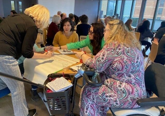 A group of women sit around a table, collaborating on a large sheet of paper during a RAID Business Plan 2026 Survey workshop. Other participants are engaged in discussions at tables in the background.