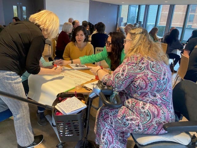 A group of women sit around a table, collaborating on a large sheet of paper during a RAID Business Plan 2026 Survey workshop. Other participants are engaged in discussions at tables in the background.