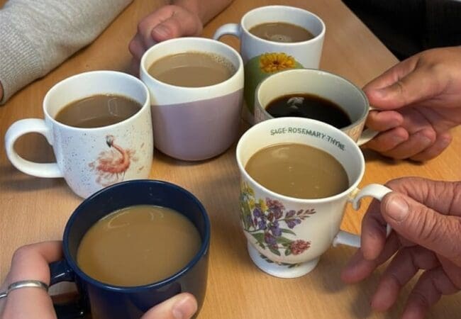 Six people sit around a table holding assorted mugs filled with tea or coffee over a wooden table -a friendly coffee session.