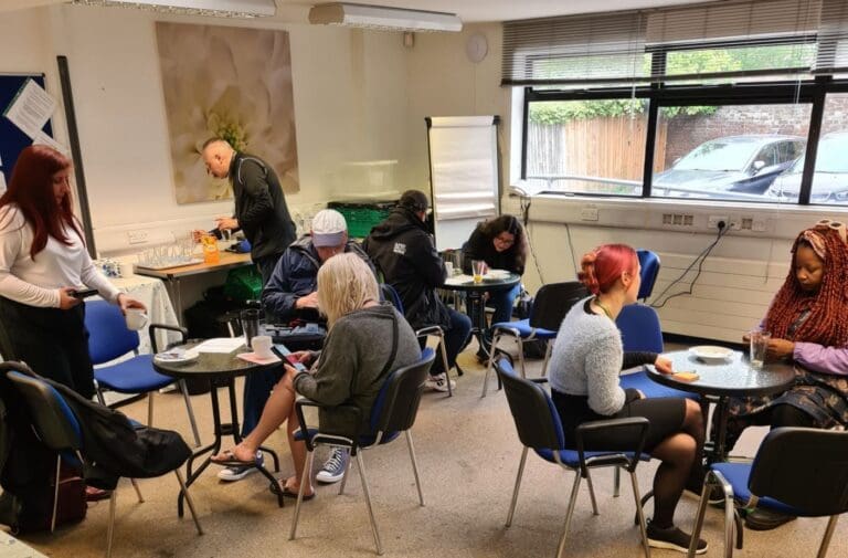 A group of people sit and stand around small tables in a brightly lit room chatting. There are large windows, blue chairs, and a flip chart in the background—a supportive space on the road to recovery.
