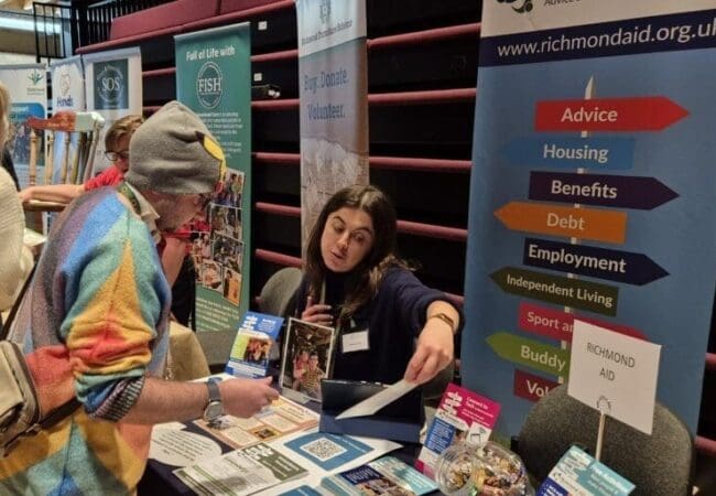 Photo of 2 people, 1 seated, at a table covered in leaflets. They are looking at and discussing one of the leaflets. A pop up banner details some of the services provided by RAID (Richmond AID).