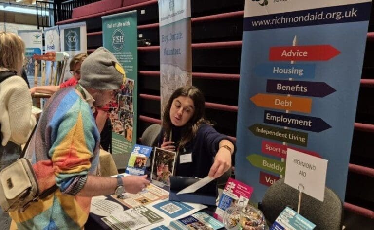 Photo of 2 people, 1 seated, at a table covered in leaflets. They are looking at and discussing one of the leaflets. A pop up banner details some of the services provided by RAID (Richmond AID).