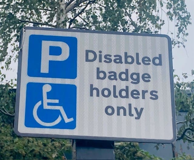 Parking sign with the wheelchair symbol and text reading Disabled badge holders only against a background of trees, clearly indicating accessible parking for Blue Badge permit holders.