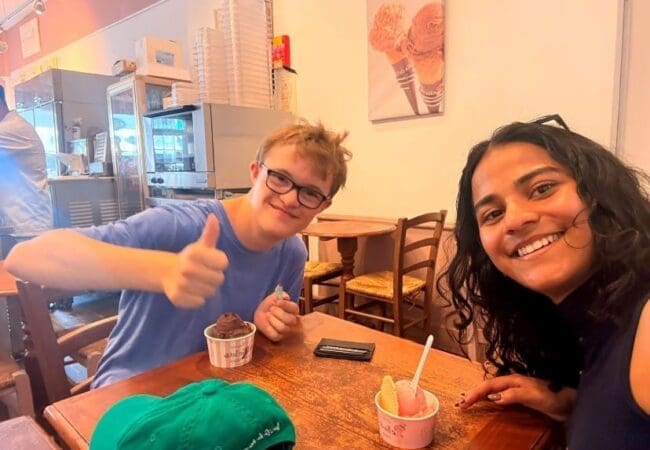 BuddyUp July 26 Newsletter Two people sit at a table in an ice cream parlour, smiling at the camera for the July Newsletter. One gives a thumbs-up while holding ice cream; the other is leaning in with a grin, perhaps celebrating a sweet end to a successful RAID. Cups of gelato and a green cap are on the table.