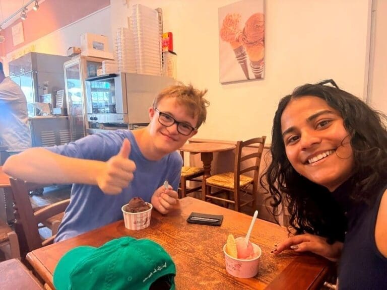 Two people sit at a table in an ice cream parlour, smiling at the camera for the July Newsletter. One gives a thumbs-up while holding ice cream; the other is leaning in with a grin, perhaps celebrating a sweet end to a successful RAID. Cups of gelato and a green cap are on the table.