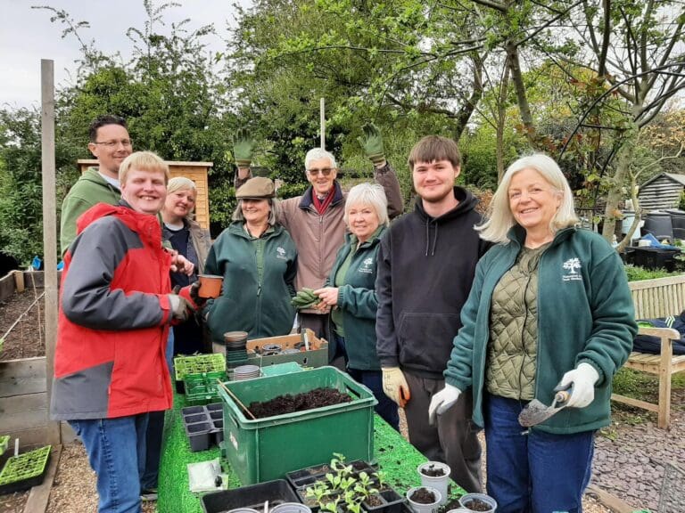 A group of people smile while gardening outdoors, standing around a table with soil, pots, and young plants. Trees and garden beds are visible in the background, highlighting the joy found in volunteering opportunities that bring communities together.