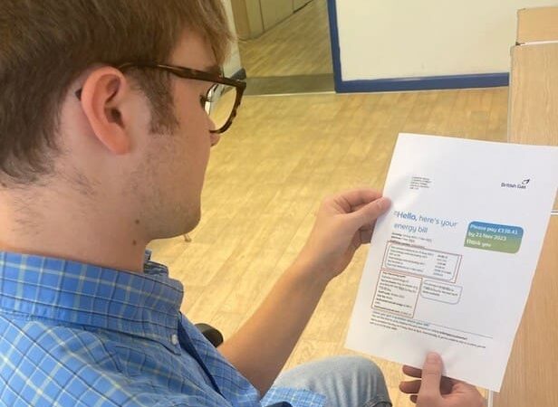 A person wearing glasses and a blue checked shirt sits at a desk, looking at an energy bill. The bill features bold text, tables, and a coloured section showing the amount due—a clear reminder of recent energy price rises.