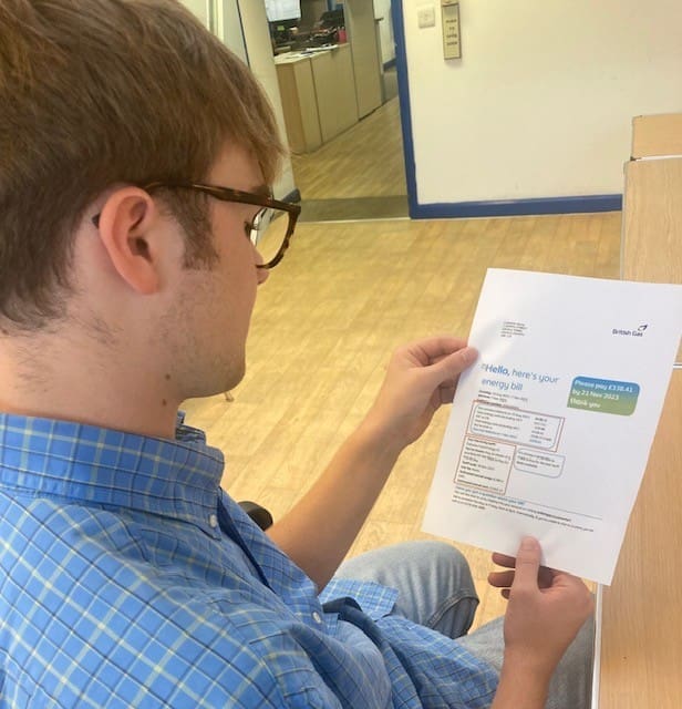 A person wearing glasses and a blue checked shirt sits at a desk, looking at an energy bill. The bill features bold text, tables, and a coloured section showing the amount due—a clear reminder of recent energy price rises.