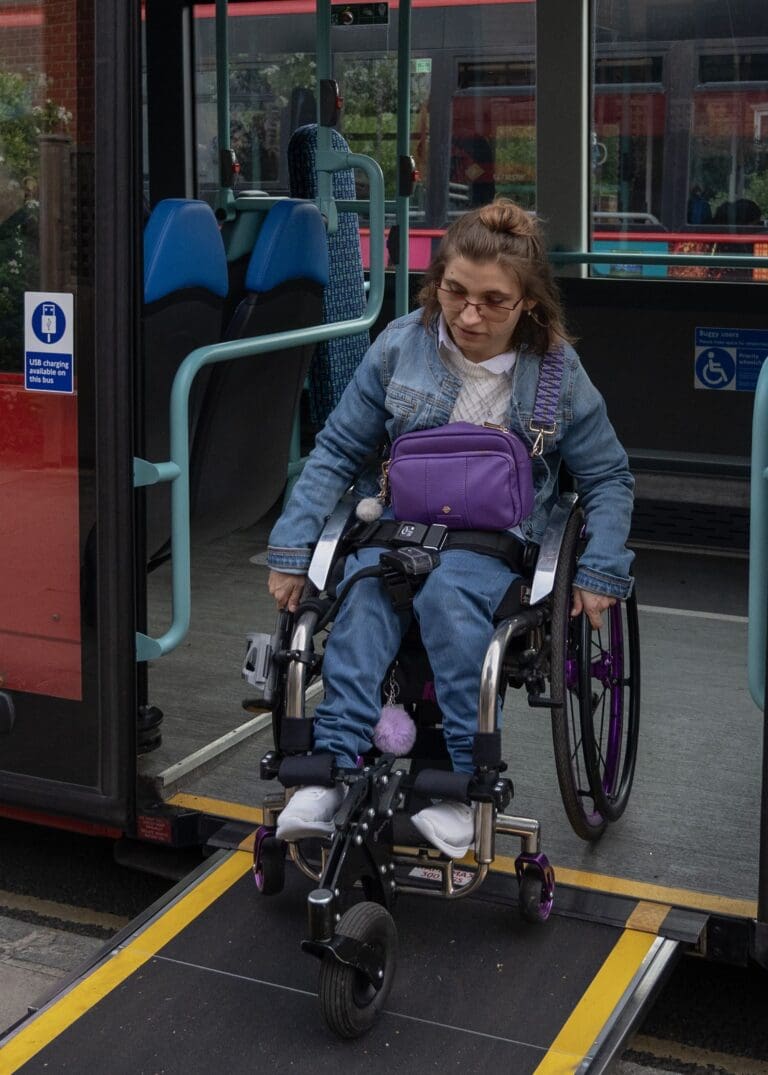 A woman using a wheelchair exits a bus via a ramp. She wears glasses, a denim jacket, and jeans, with a purple bag on her lap. The bus has blue seats and accessibility signs, reflecting the government's commitment to supporting disabled people through free bus travel initiatives.