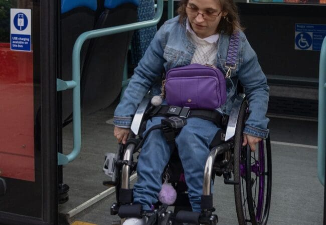 A woman using a wheelchair exits a bus via a ramp. She wears glasses, a denim jacket, and jeans, with a purple bag on her lap. The bus has blue seats and accessibility signs, reflecting the government's commitment to supporting disabled people through free bus travel initiatives.