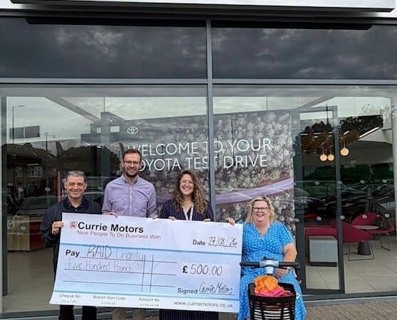Four people, one using a mobility scooter, stand outside Currie Motors holding a large £500 charity cheque made out to RADI Cornwall. A Toyota dealership sign and vehicles are visible in the background, as the group gathers to express their donation thanks for this generous contribution.