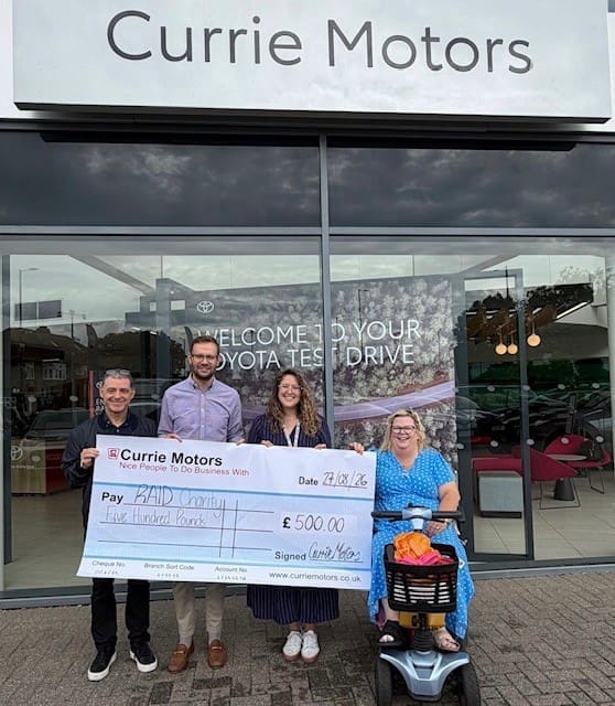 Four people, one using a mobility scooter, stand outside Currie Motors holding a large £500 charity cheque made out to RADI Cornwall. A Toyota dealership sign and vehicles are visible in the background, as the group gathers to express their donation thanks for this generous contribution.