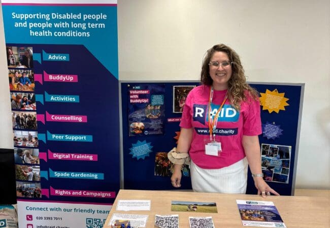 A woman in a pink RAID shirt stands behind a table with leaflets, pens, and forms, inviting BuddyUp Volunteers to join community service. A RAID disability support banner and information display board are beside and behind her.