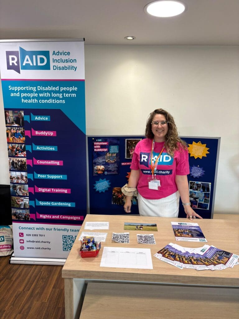 A woman in a pink RAID shirt stands behind a table with leaflets, pens, and forms, inviting BuddyUp Volunteers to join community service. A RAID disability support banner and information display board are beside and behind her.