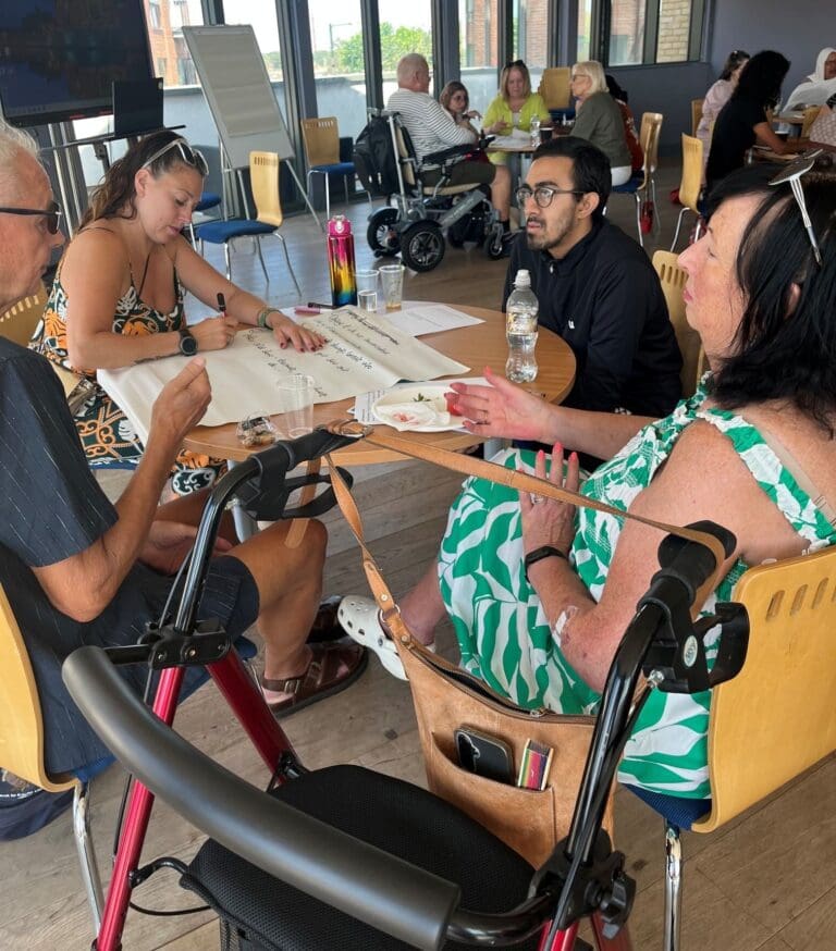 Four people, including two with mobility aids, sit around a table writing on a large sheet of paper in a bright, open room with other groups in the background, engaged in discussion and collaboration as part of the September Newsletter feature on RAID initiatives.